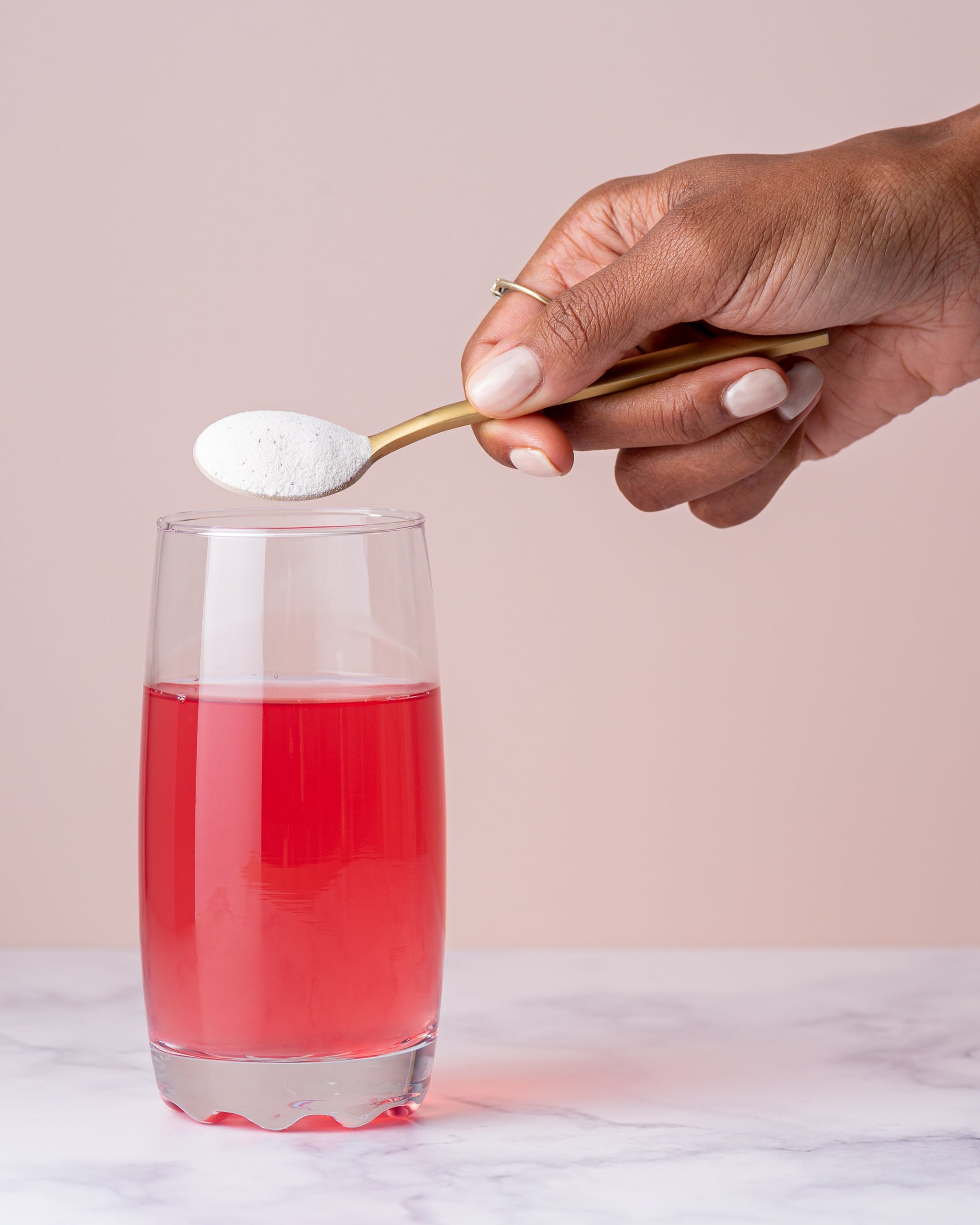 Cuillère dorée de poudre Bend Beauty Collagène marin + Co-facteurs tenue au-dessus d’un verre de boisson rose