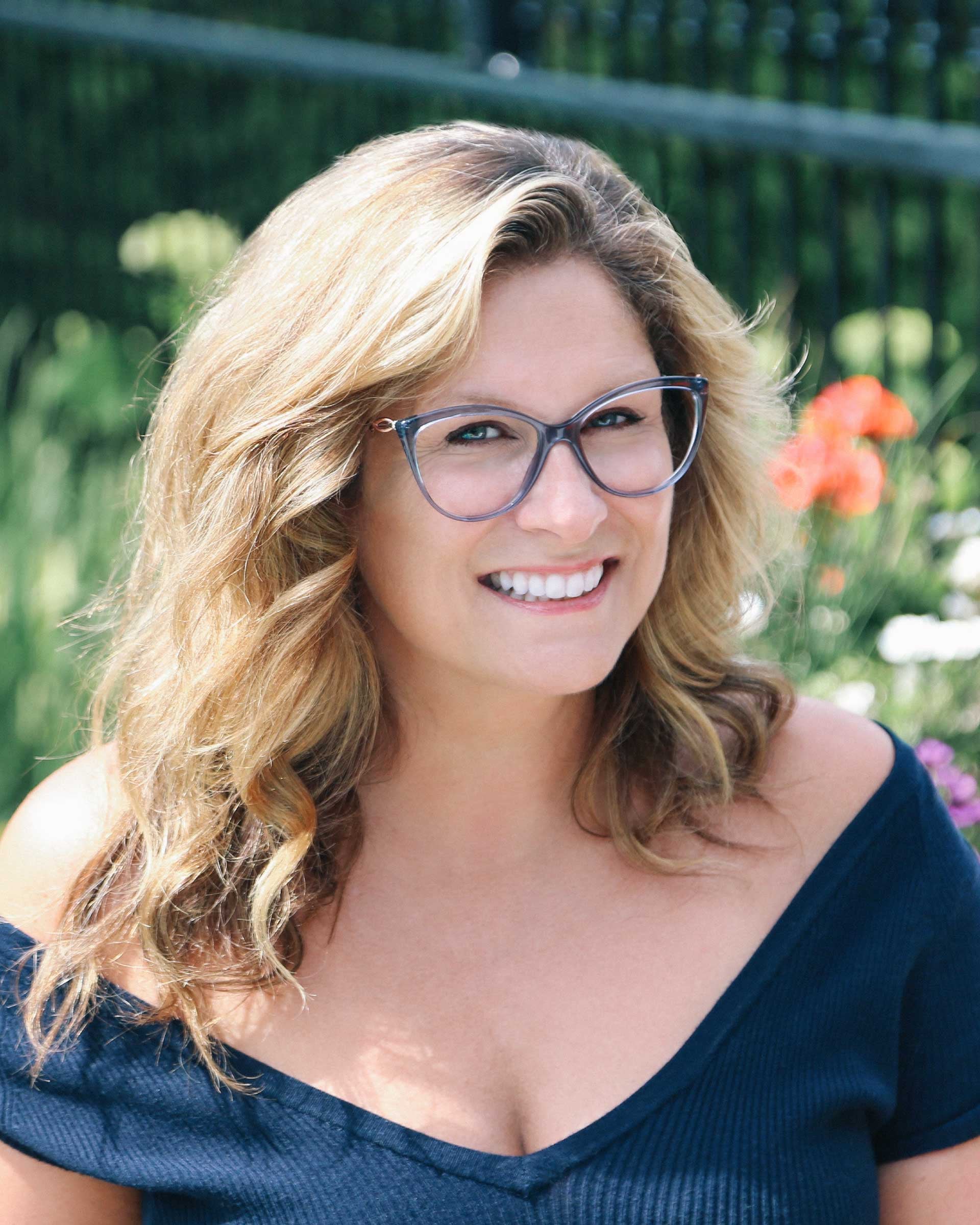 Saskia Thuot, ambassadrice d'IRIS, sourit dans un jardin fleuri, lunettes à monture bleu-gris translucide et haut marine aux épaules dénudées