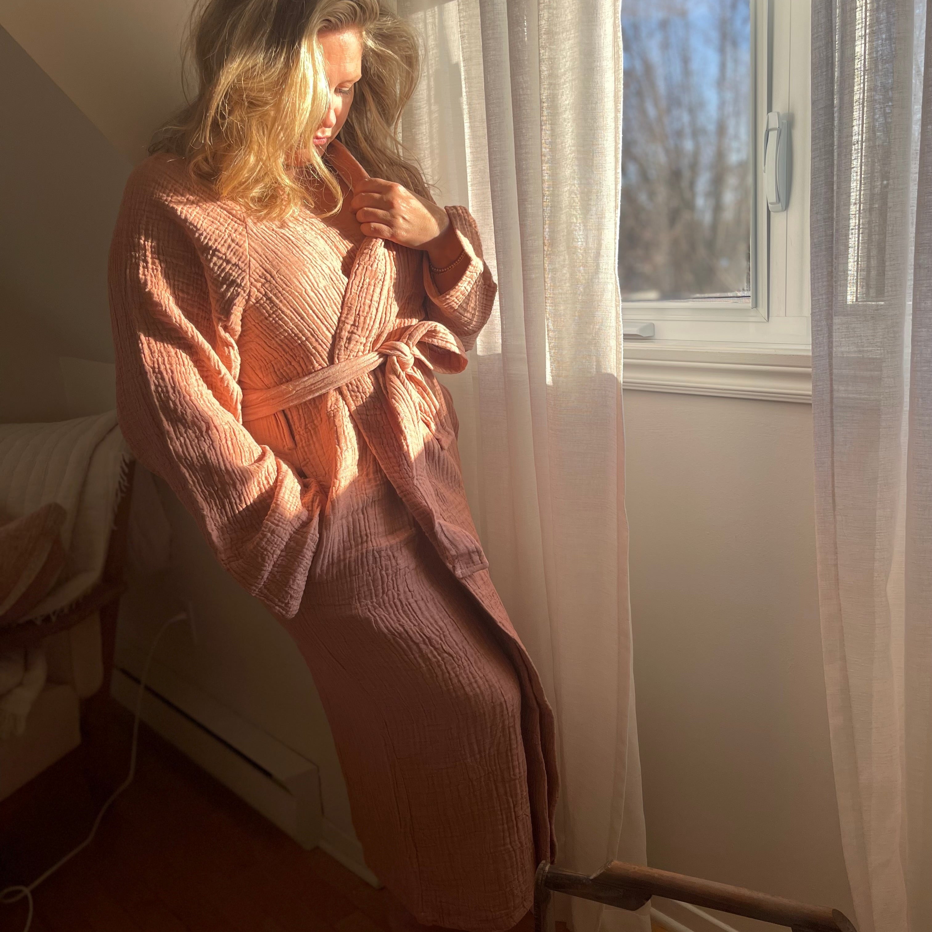 Woman standing by a window in soft morning light, wearing the Bella Bathrobe – Terracotta