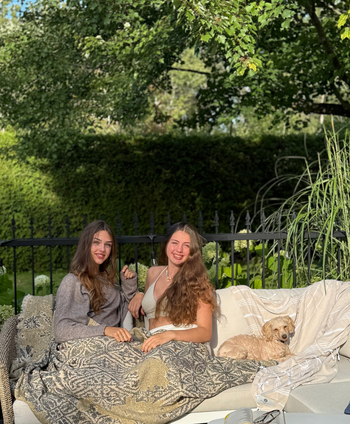Women smiling wrapped in the Georgie throw made of natural cotton, sitting outdoors in the sun. Soft and chic throw from Balmy’s collection.