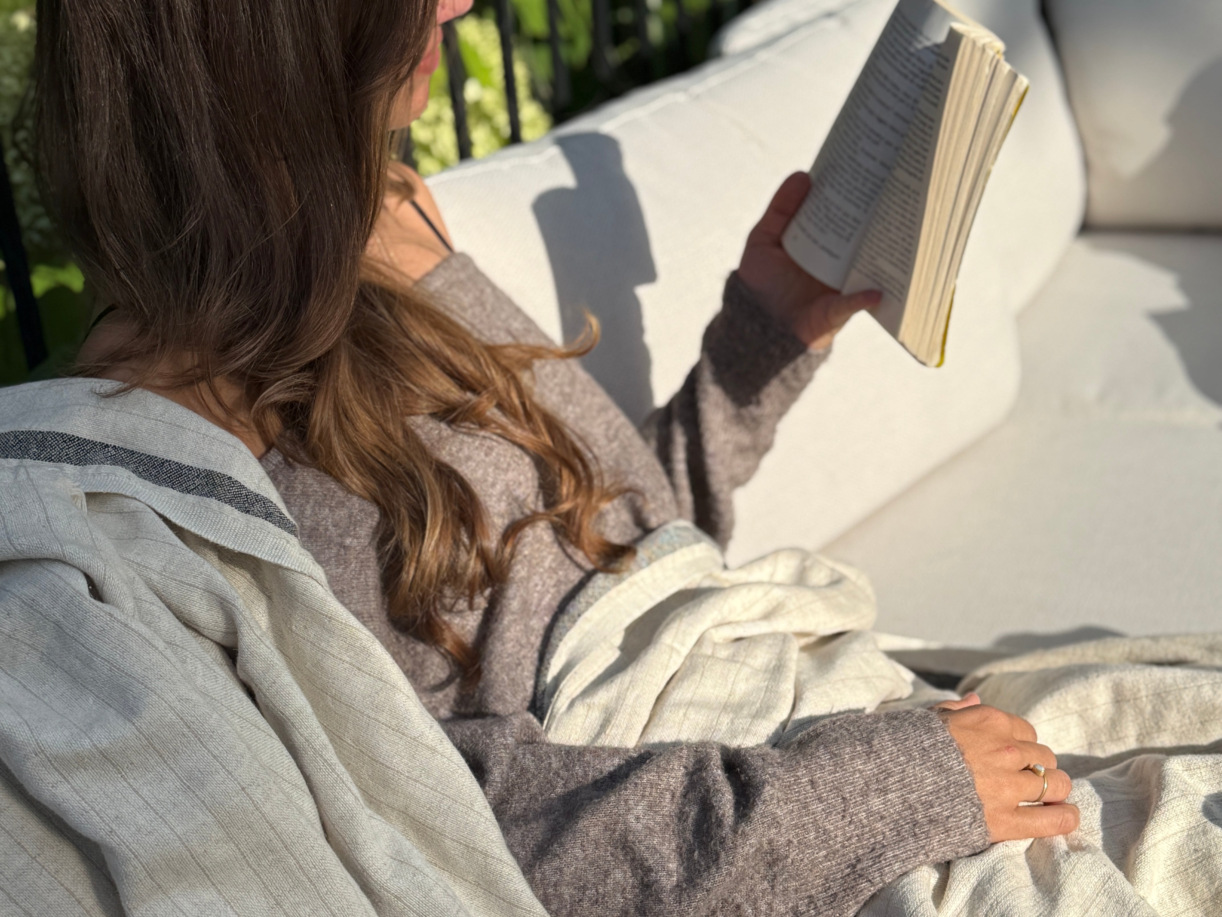 Woman reading outside wrapped in a natural linen Terri throw, sunlight and greenery in the background.