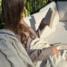 Woman reading outside wrapped in a natural linen Terri throw, sunlight and greenery in the background.