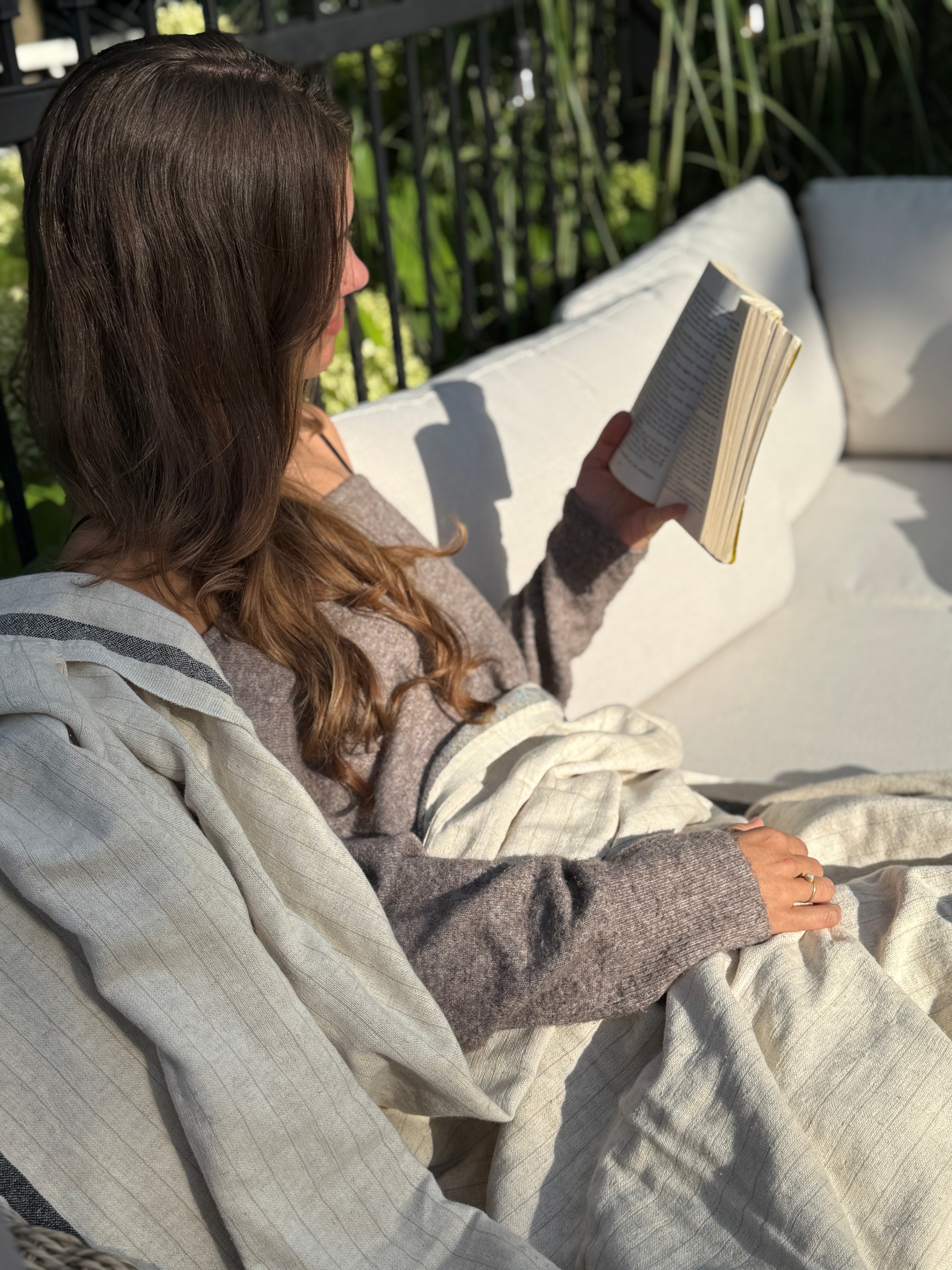 Woman reading outside wrapped in a natural linen Terri throw, sunlight and greenery in the background.