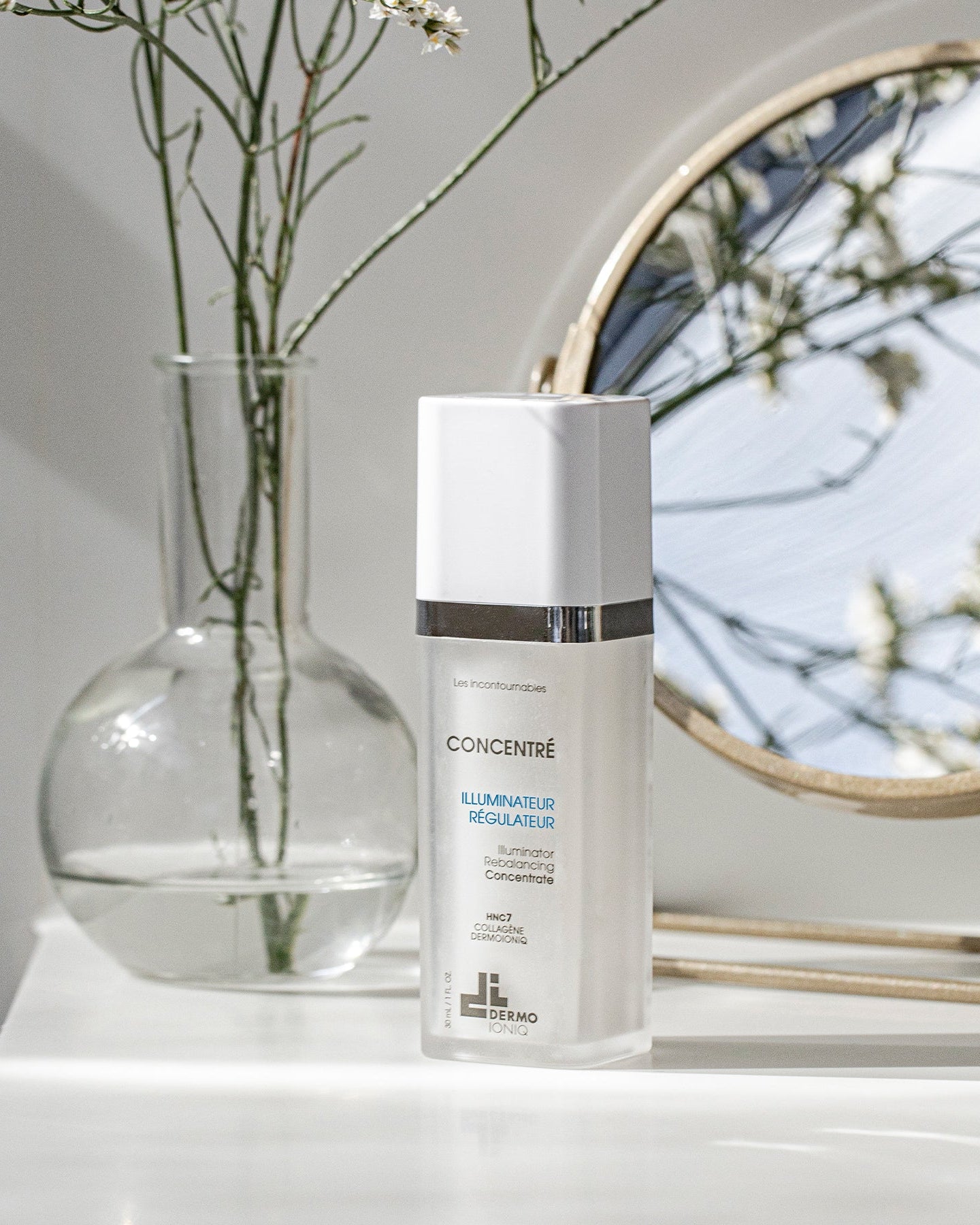 Frosted Dermo Ioniq Illuminator Rebalancing Concentrate bottle on a white shelf beside a glass vase and a round mirror