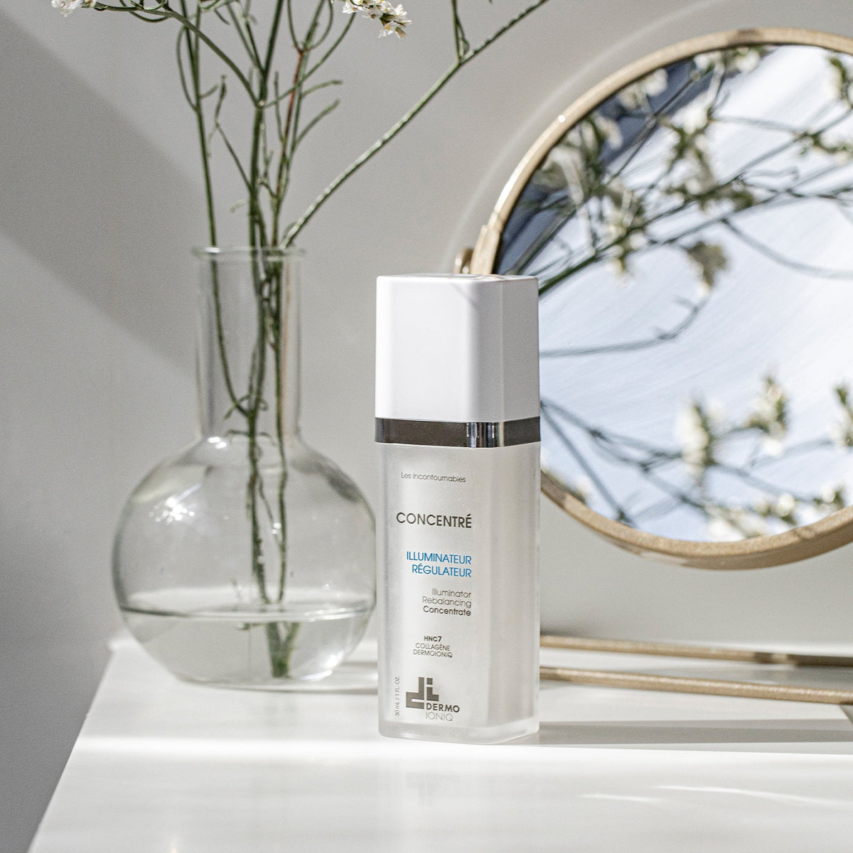 Frosted Dermo Ioniq Illuminator Rebalancing Concentrate bottle on a white shelf beside a glass vase and a round mirror