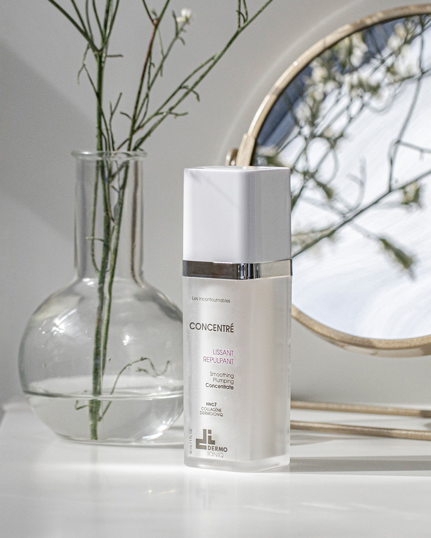 Frosted Dermo Ioniq Smoothing Plumping Concentrate bottle on a white shelf beside a glass vase and a round mirror