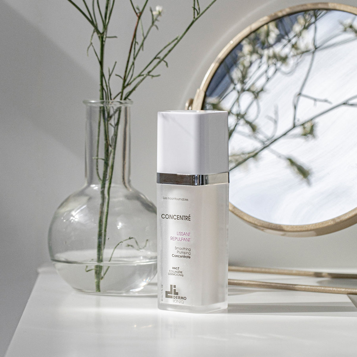 Frosted Dermo Ioniq Smoothing Plumping Concentrate bottle on a white shelf beside a glass vase and a round mirror