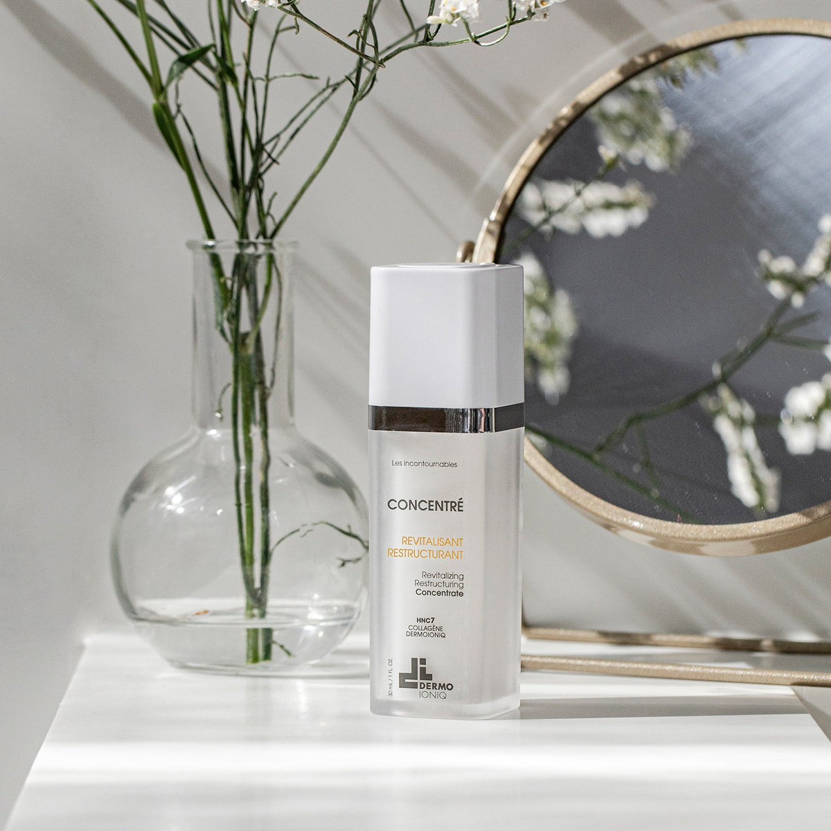 Frosted Dermo Ioniq Revitalizing Restructuring Concentrate bottle on a white shelf beside a glass vase and a mirror
