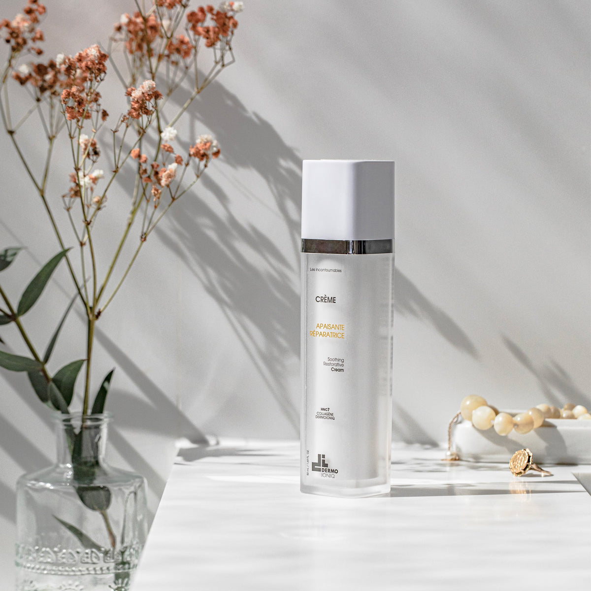 Flacon givré Dermo Ioniq Crème Apaisante Réparatrice sur une tablette blanche, près d’un vase de fleurs séchées