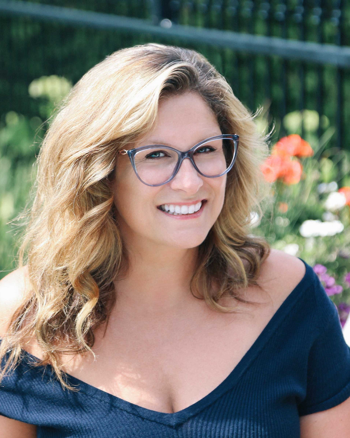 Saskia Thuot, ambassadrice d'IRIS, sourit dans un jardin fleuri, lunettes à monture bleu-gris translucide et haut marine aux épaules dénudées