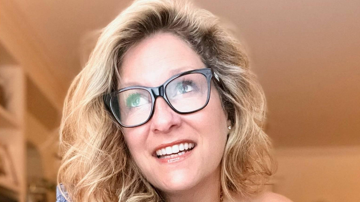 Saskia Thuot sourit en levant les yeux, lunettes à monture noire et cheveux blonds ondulés, dans un intérieur chaleureux