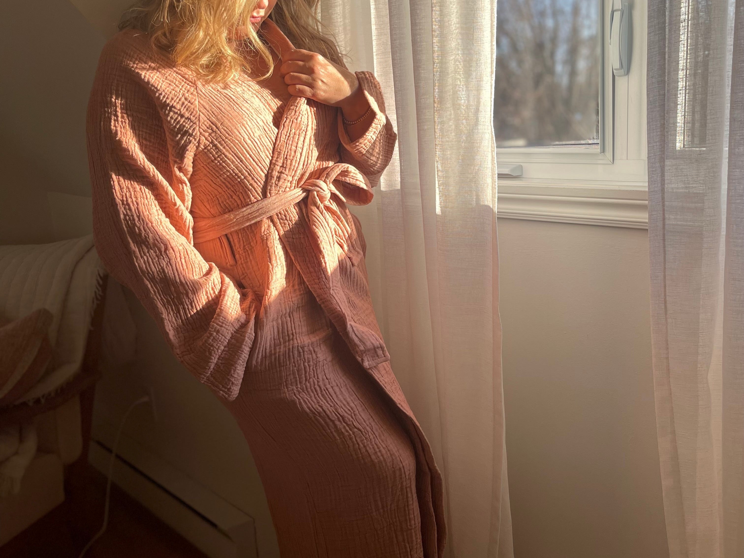 Woman standing by a window in soft morning light, wearing the Bella Bathrobe – Terracotta

