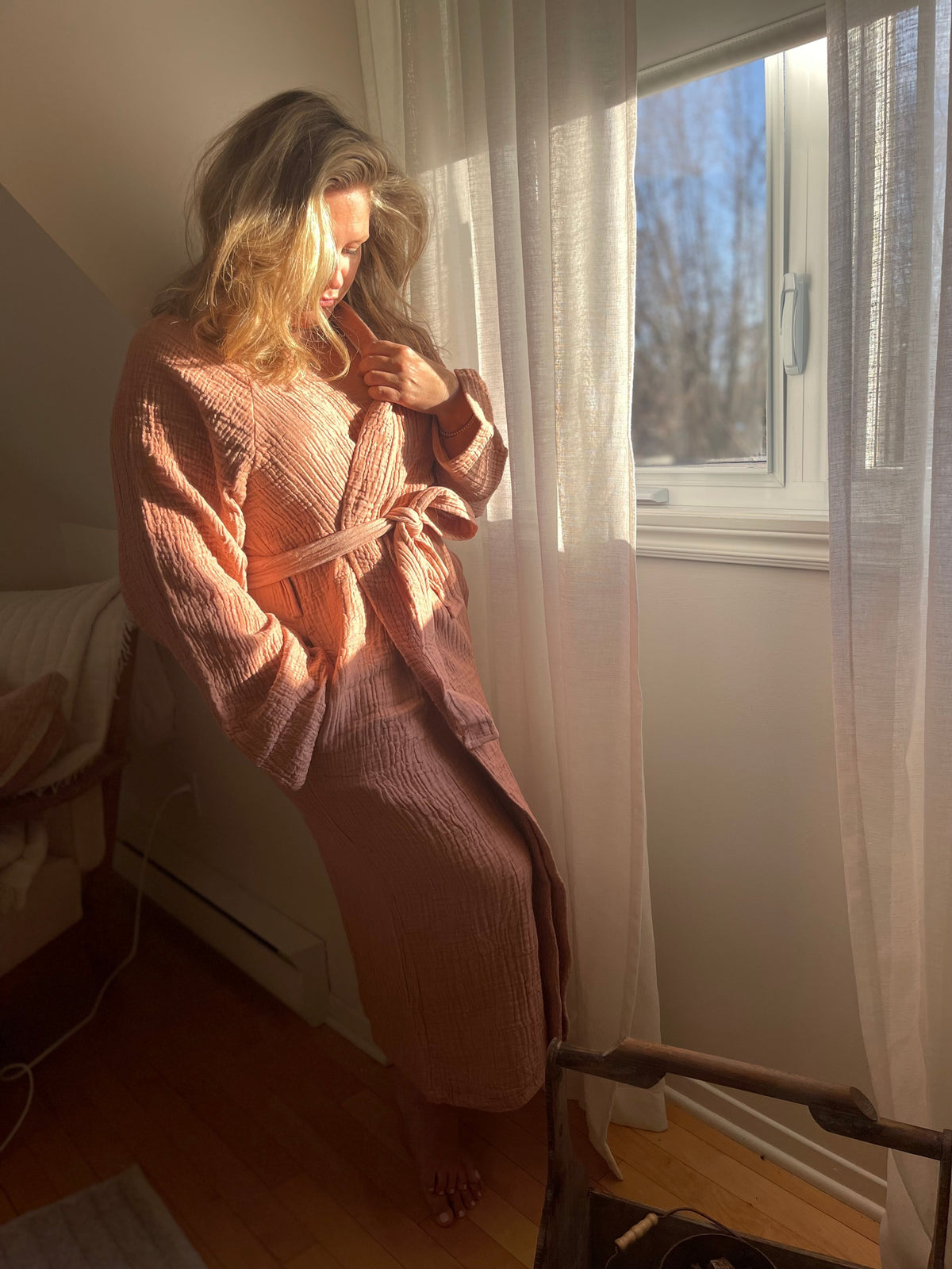 Femme debout près d'une fenêtre, dans une douce lumière, vêtue du peignoir BELLA terre cuite