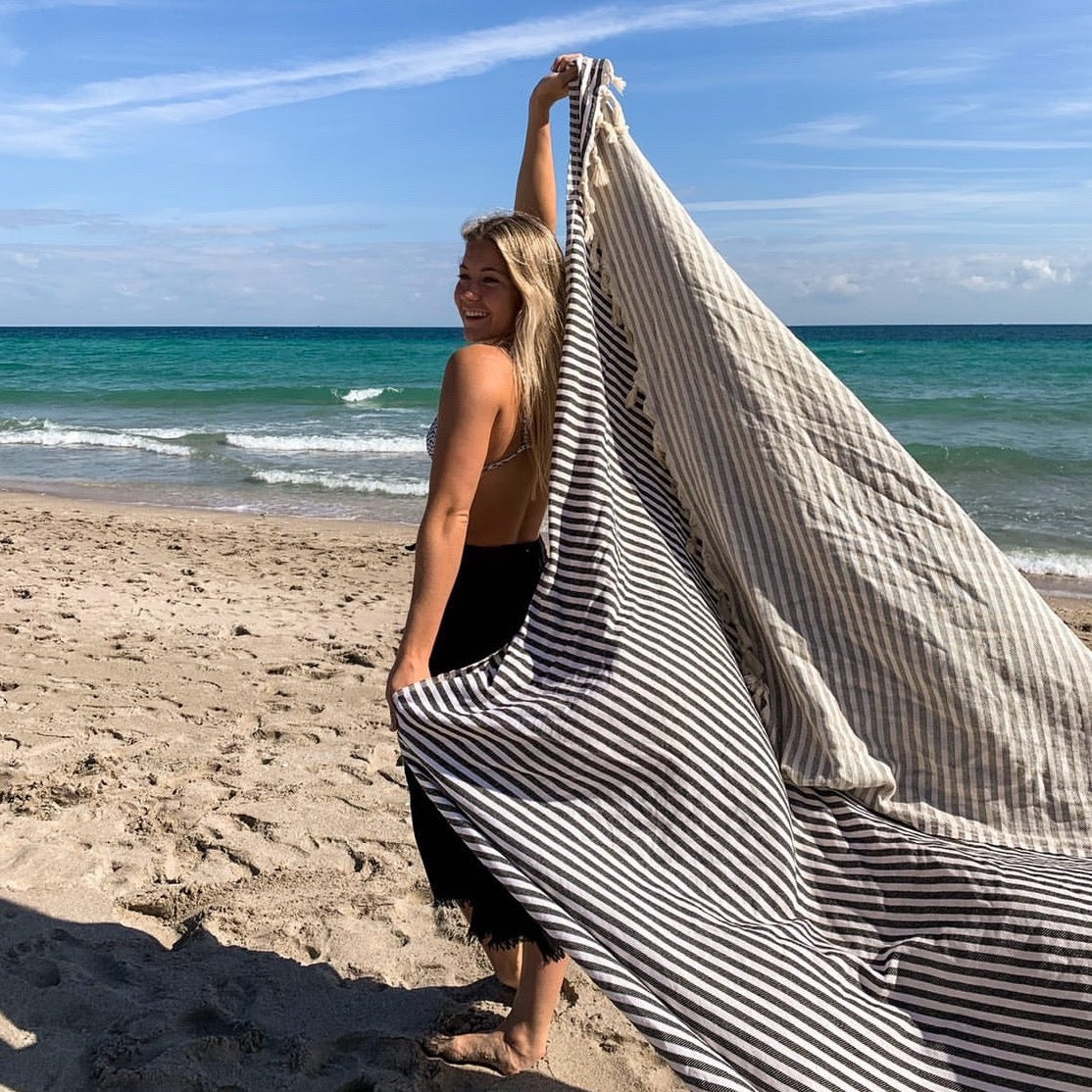 Smiling woman holding a black and white striped Balmy Serena XL Turkish towel on a sunny beach, with turquoise ocean waves in the background.